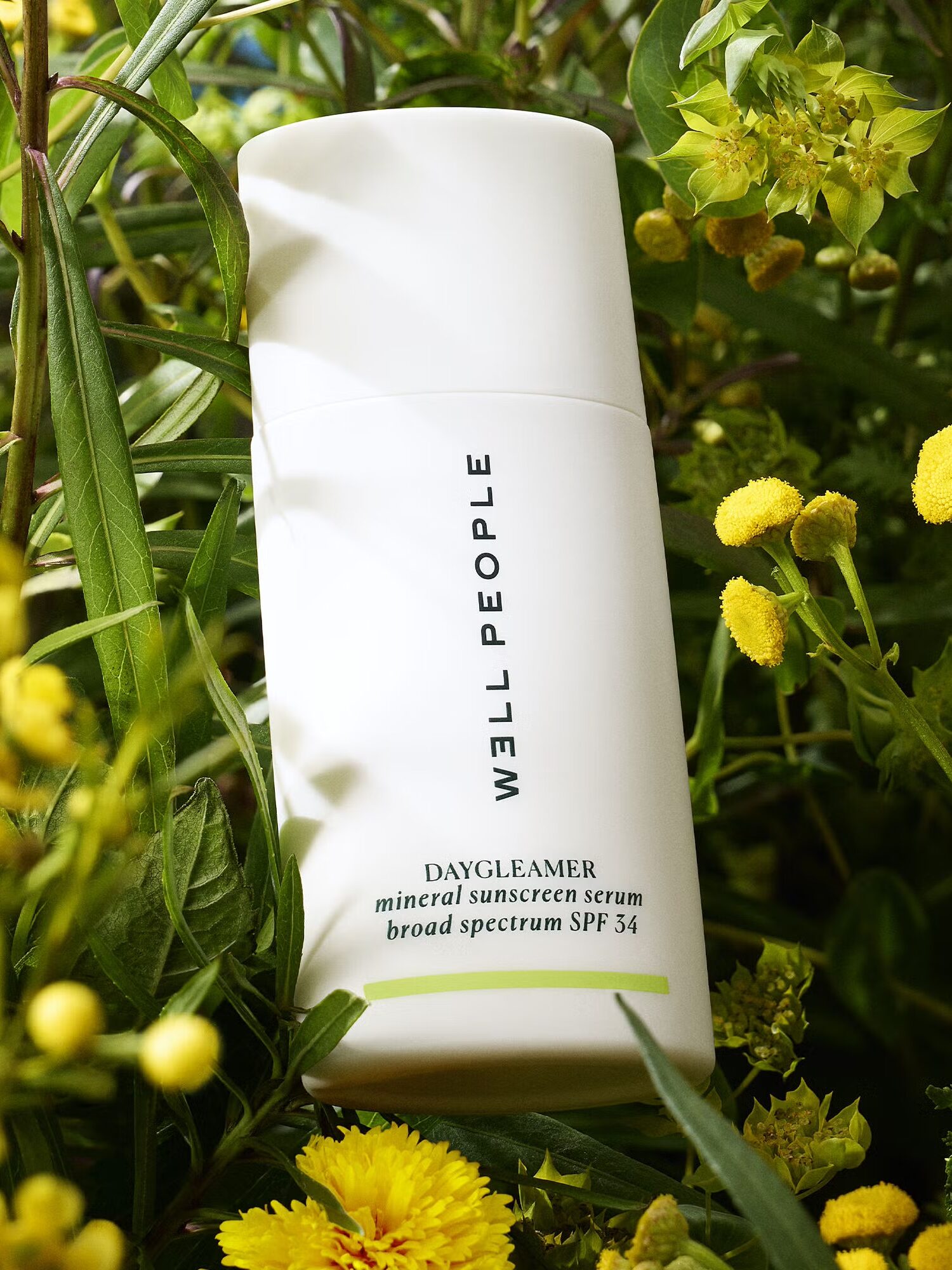 Bottle of Well People Daydreamer mineral sunscreen serum SPF 34 surrounded by green leaves and yellow flowers.