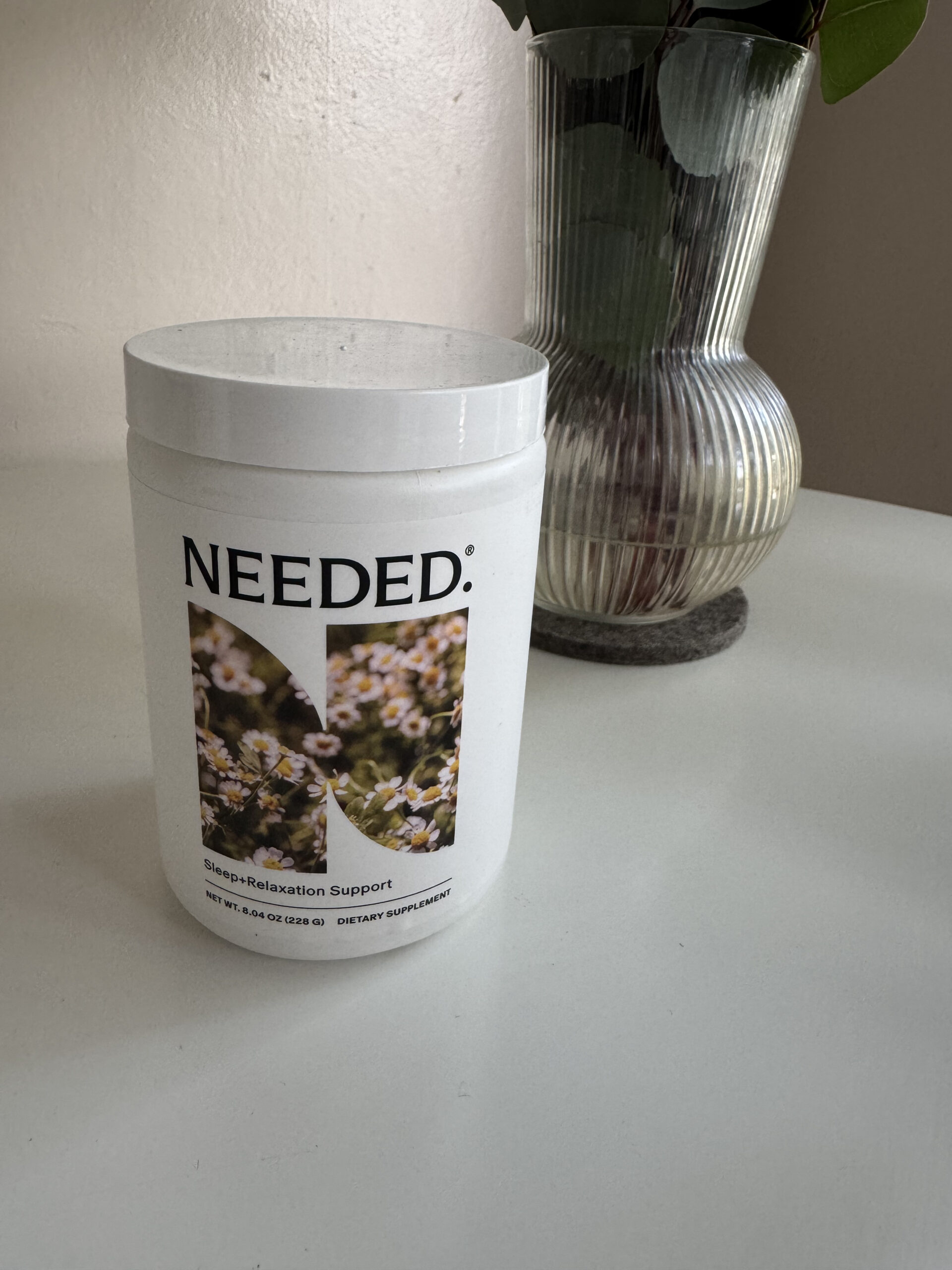 A white container labeled "NEEDED. Sleep + Relaxation Support" is placed on a white surface next to a ribbed glass vase with leaves.