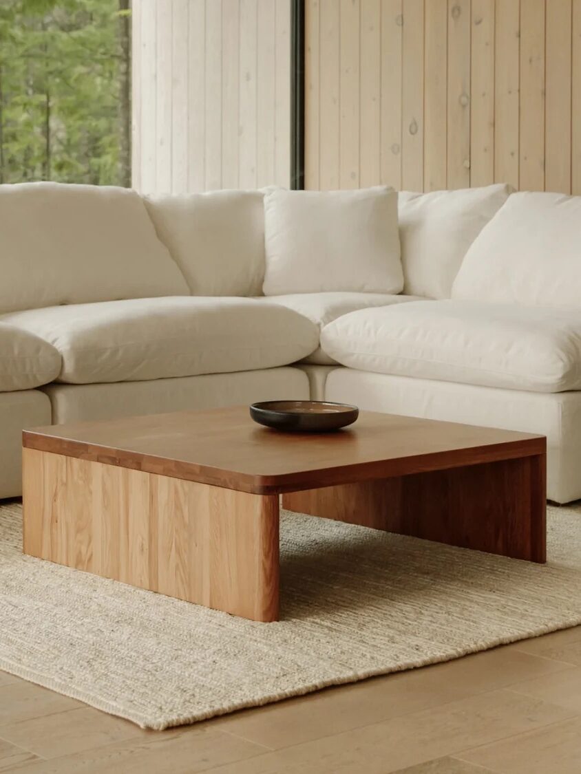 A beige sectional sofa sits on a light rug with a wooden coffee table in front. Large windows reveal a green outdoor view. The room has wooden paneled walls and hardwood flooring.