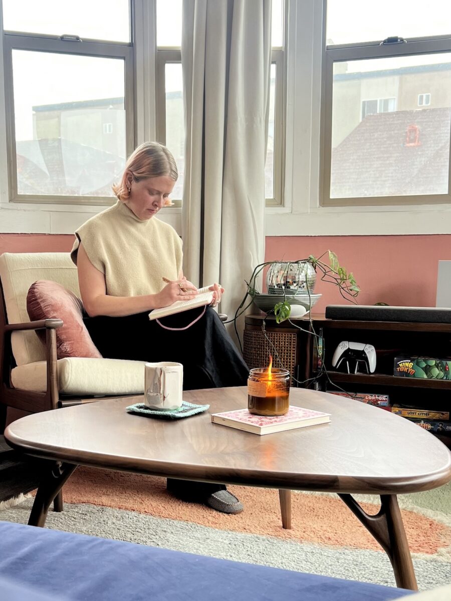 Person sitting in a chair, writing in a notebook, with a candle on a coffee table and various items in the background.