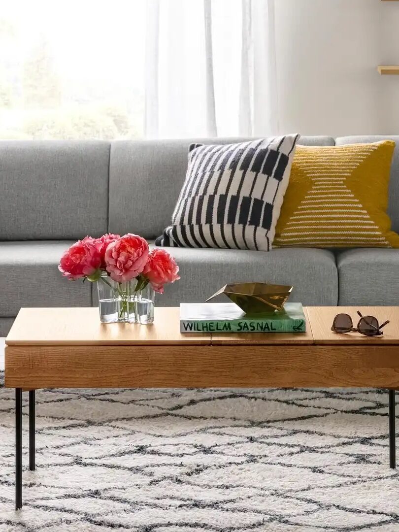 A modern living room with a gray sofa, patterned pillows, and a wooden coffee table with books and flowers. A cart with bottles is on the left, and a geometric rug covers the floor.