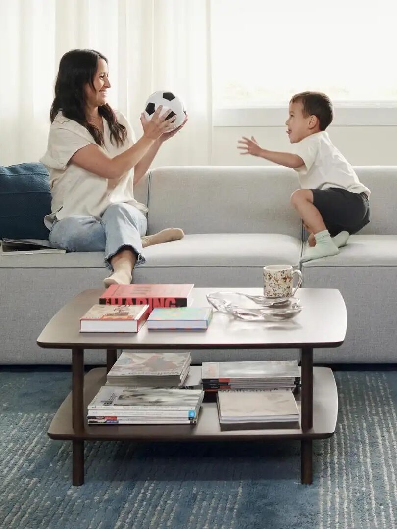 A family of three playing with a soccer ball on a gray sofa in a bright living room. The woman tosses the ball to the child, and the man watches. A coffee table with books is in front.