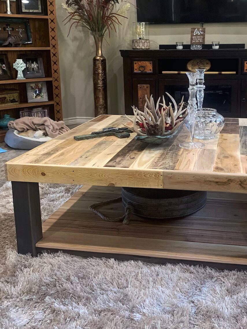 A square wooden coffee table with a rustic design on a beige shag rug. It has a lower shelf with a coiled rope and is decorated with a glass decanter and antler centerpiece.