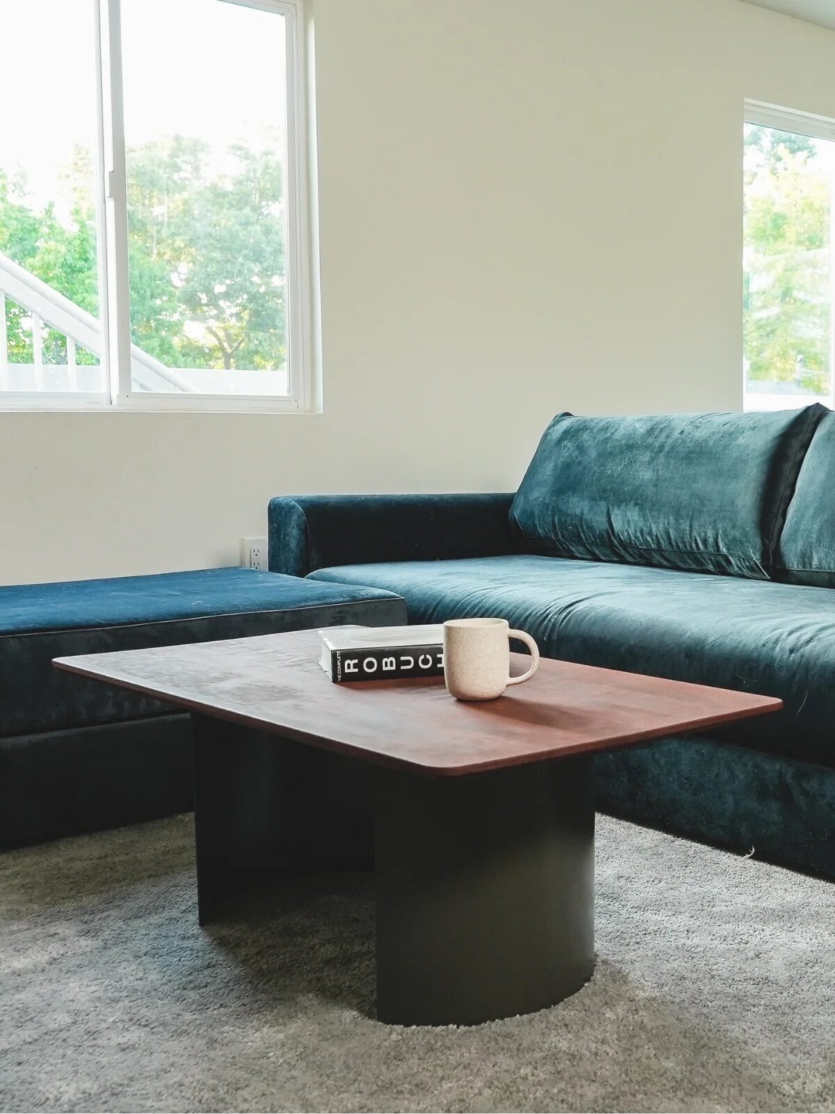 A living room with a dark green velvet sofa, matching ottoman, and wooden coffee table on a gray rug, with large windows in the background.