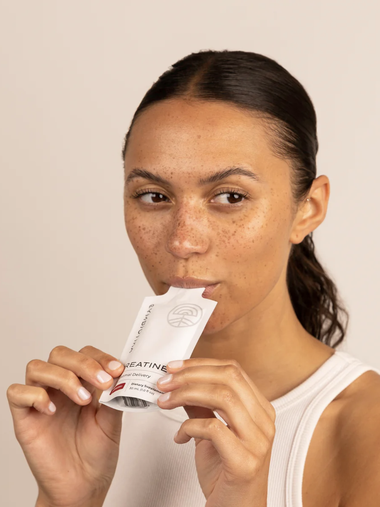 A person with long hair and freckles is sipping from a sachet labeled "Creatine." They are wearing a white tank top against a plain background.