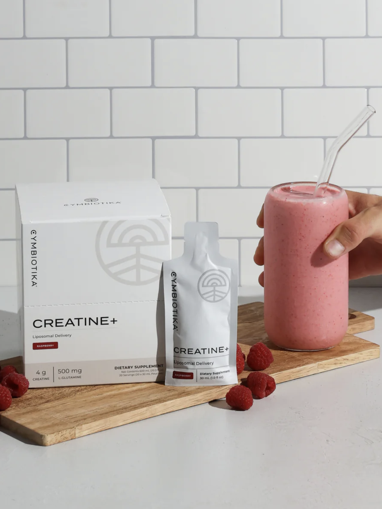 A hand holds a pink smoothie in a glass with a clear straw next to a creatine supplement box and packet on a wooden board, surrounded by raspberries against a tile background.