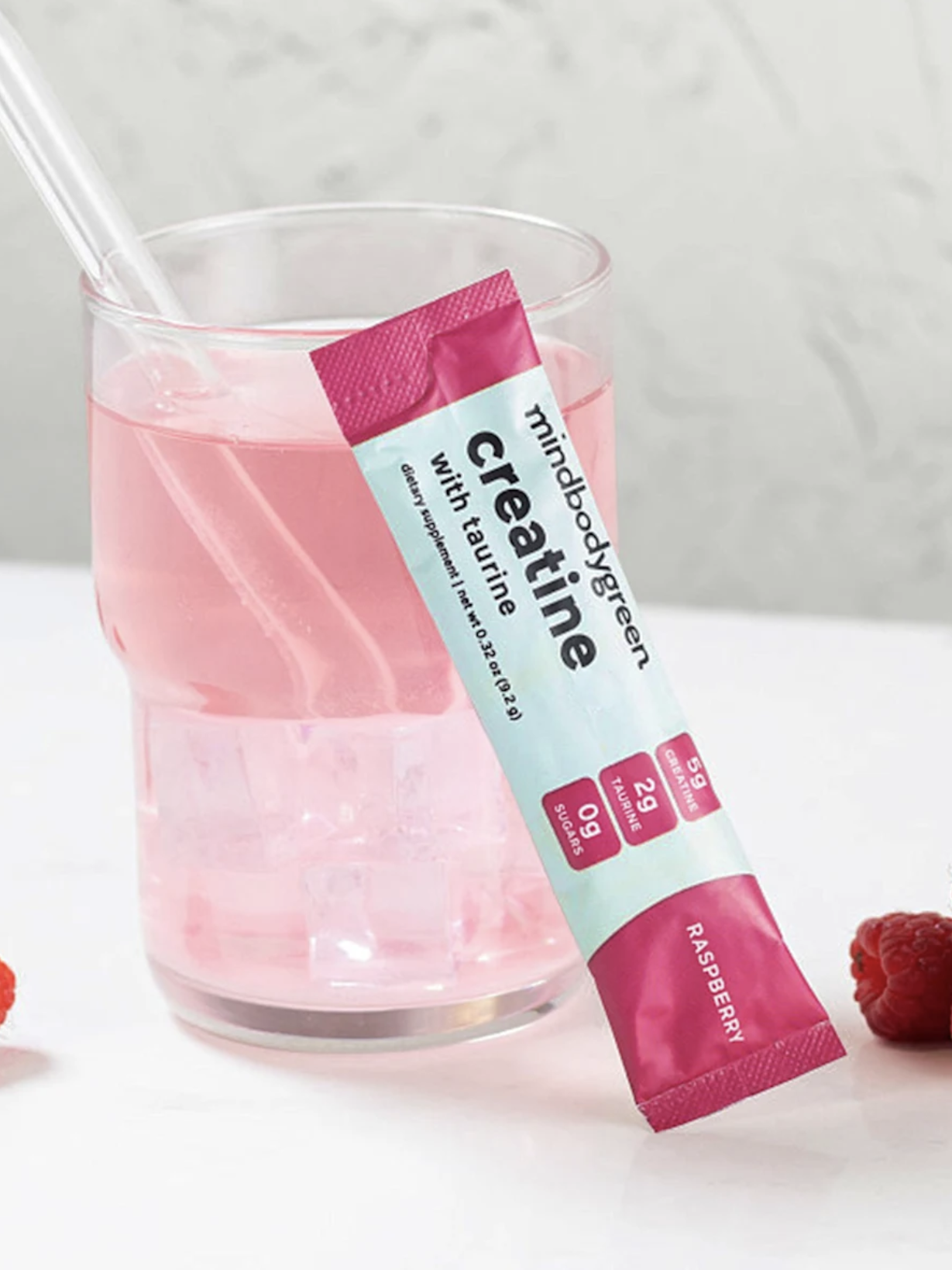 A glass of pink raspberry-flavored creatine drink with a clear straw, placed beside a creatine packet and fresh raspberries on a white surface.