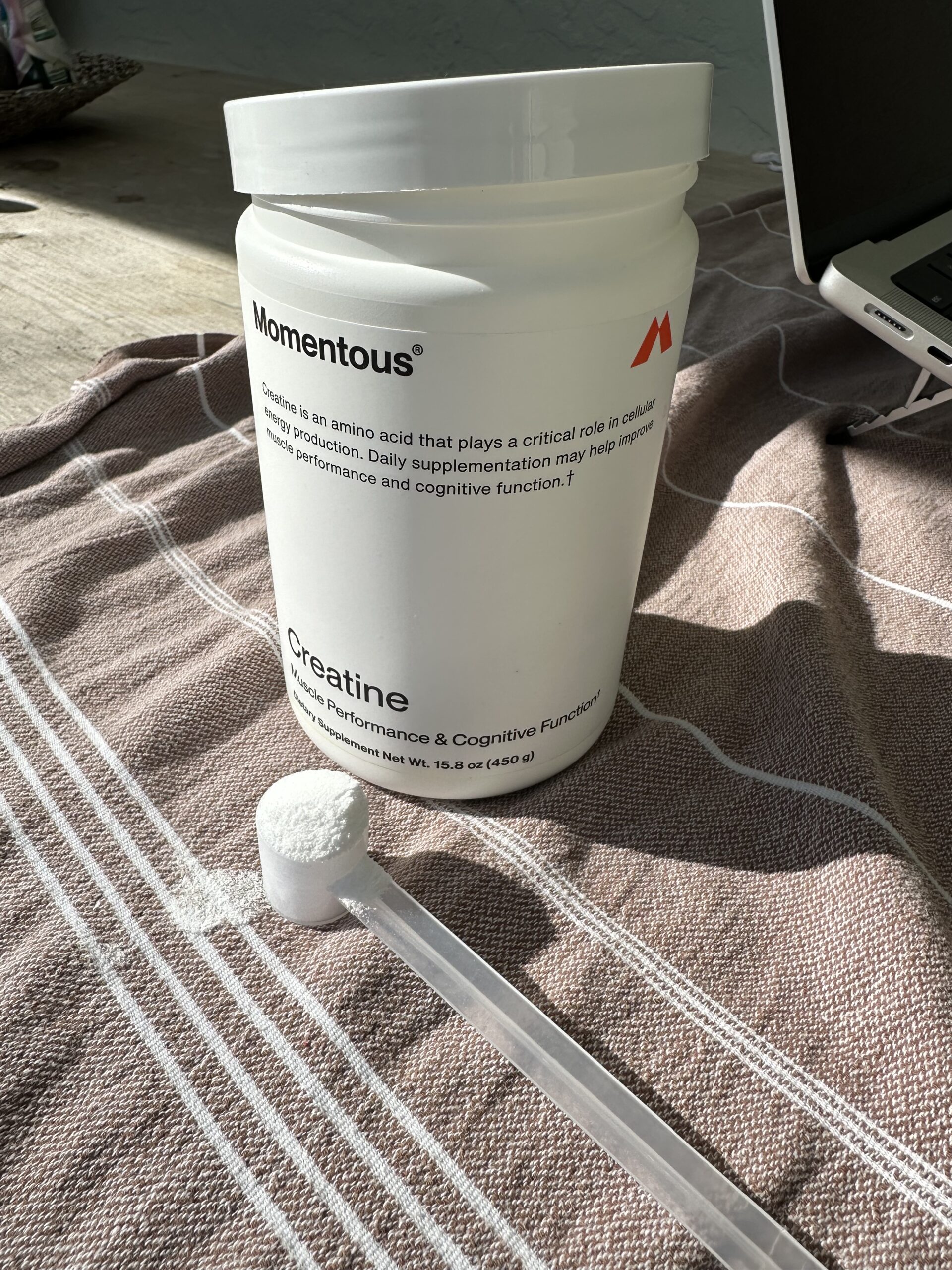 A container of Momentous creatine with a scoop of powder on a striped cloth near a laptop.