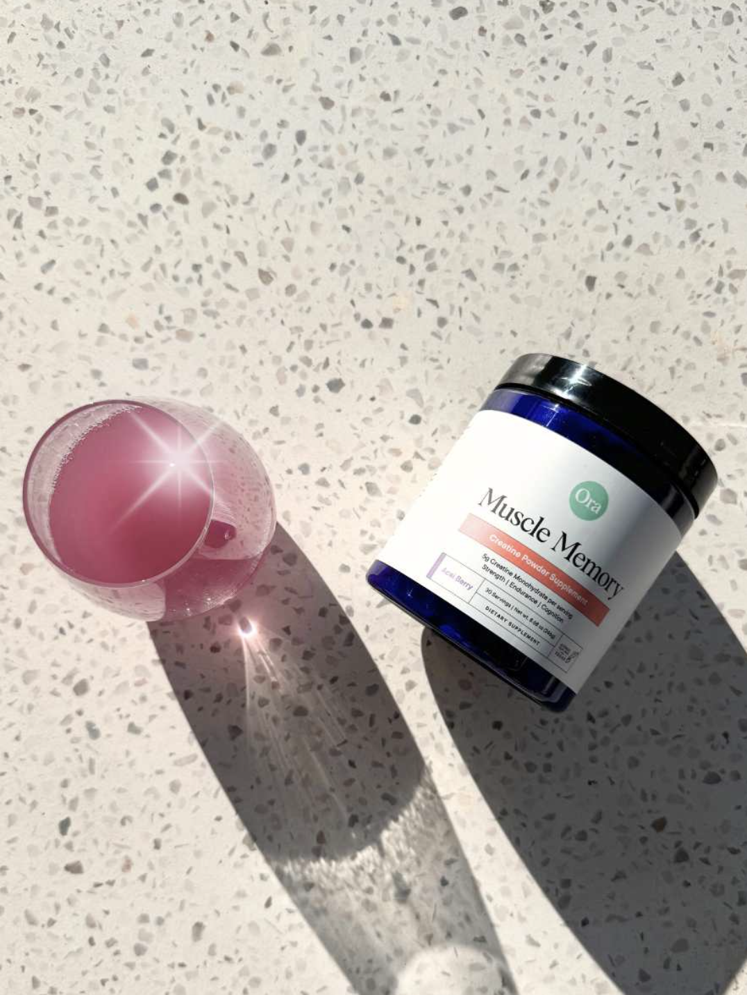 A glass of pink liquid beside a dark blue jar labeled "Muscle Memory." Both are placed on a speckled countertop with bright sunlight casting shadows.