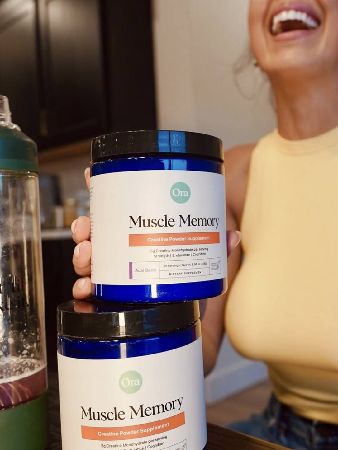 A woman in a yellow top holds a jar labeled "Muscle Memory." Another jar and a green drink bottle are on the table.