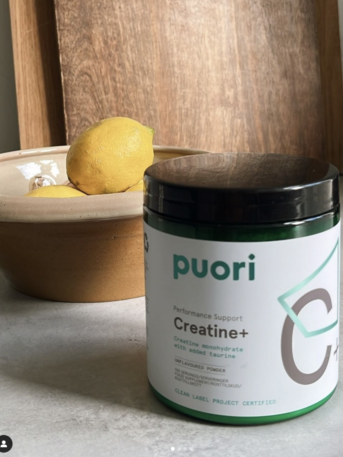Jar of Puori Creatine+ on a countertop beside a bowl with lemons; wooden boards in the background.