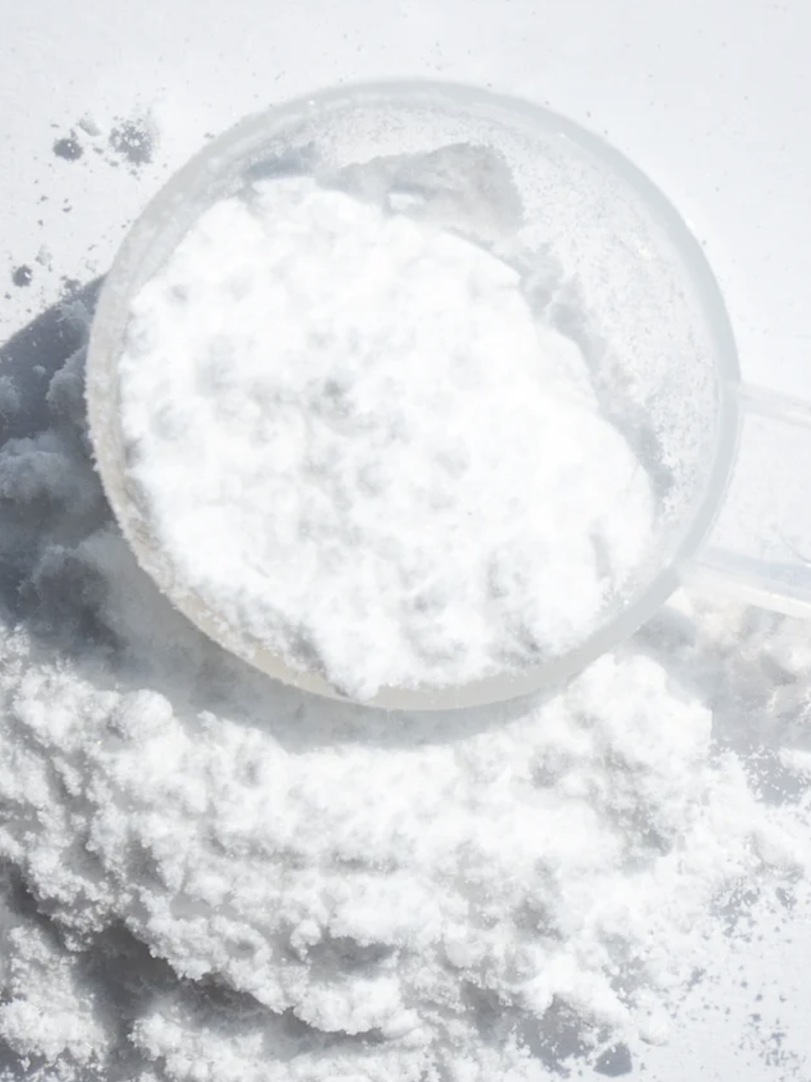 A clear plastic scoop filled with white powder rests on a small pile of the same powder, against a white background.