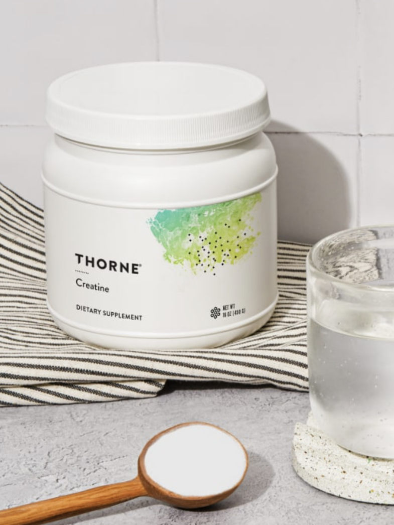 Container of Thorne creatine supplement with a wooden spoon of powder beside it and a glass of water on a coaster in the foreground.