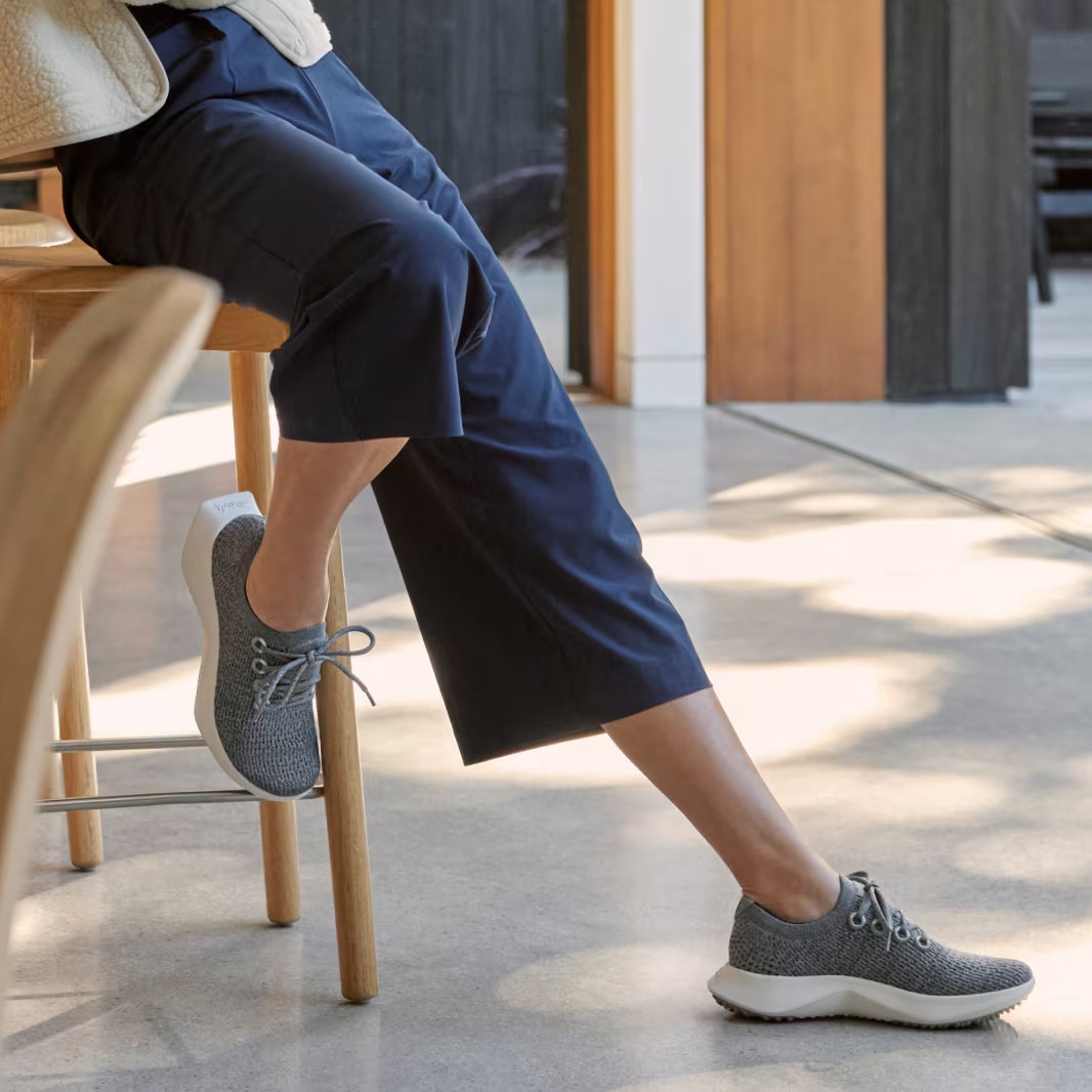 Person sitting on a stool, wearing dark pants and gray sneakers, with legs crossed in a sunlit room.