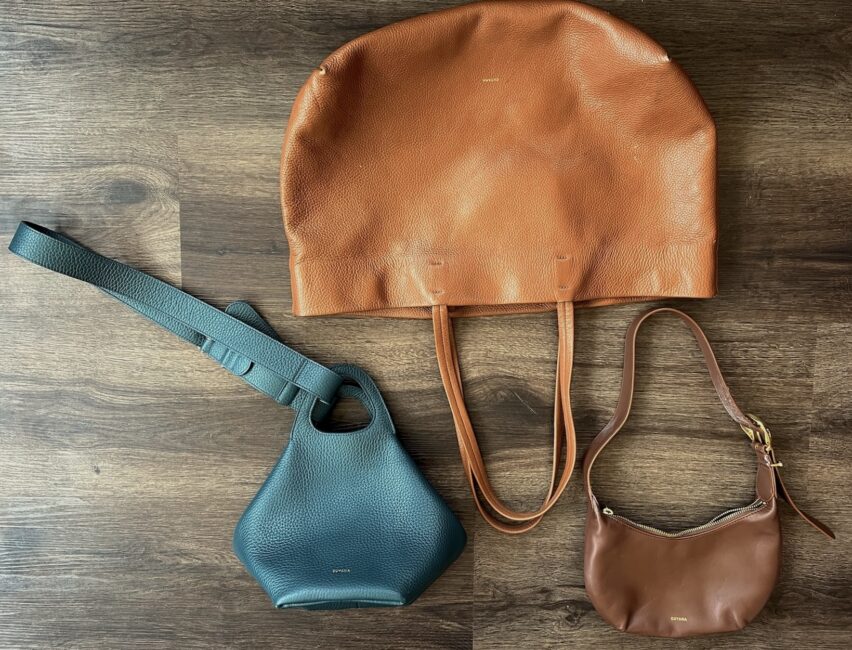 Three leather handbags—one large tan tote, one teal crossbody, and one small brown shoulder bag—are arranged on a wooden surface, creating an elegant scene worthy of a cuyana review.