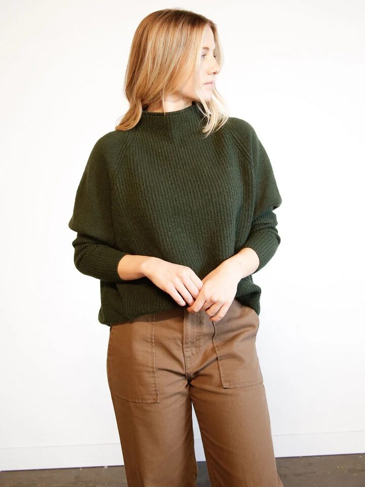 Person with long hair wearing a dark green sweater and brown pants, standing against a plain white background.