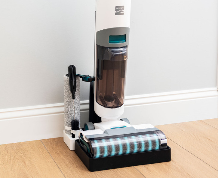 Cordless vacuum cleaner with a detachable roller is placed in its charging and cleaning dock on a wooden floor near a white wall.