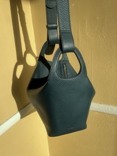 A dark green, textured leather handbag with wide handles hangs against a yellow wall, casting a shadow in the sunlight—perfect for anyone seeking quality after reading a glowing Cuyana review.