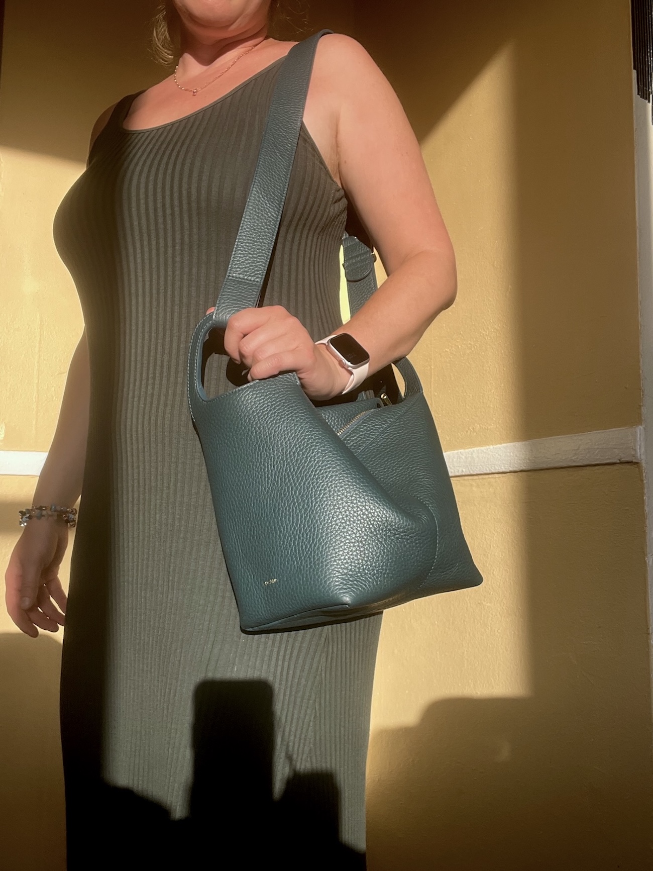 A person in a sleeveless, ribbed, dark dress stands indoors holding a textured green handbag, wearing a watch and bracelet, as sunlight casts shadows on the wall—a perfect scene for a Cuyana review.