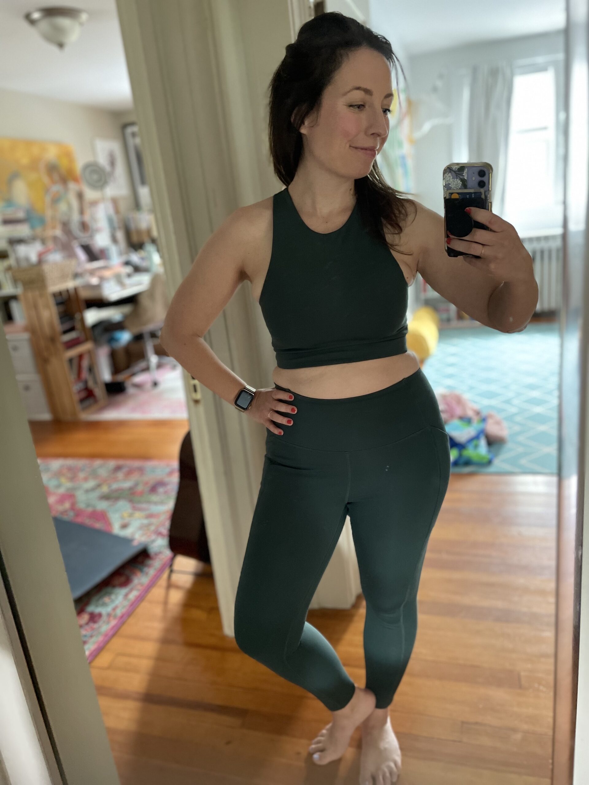 Woman in dark green athletic wear takes a mirror selfie in a well-lit room with wooden floors and various household items in the background.