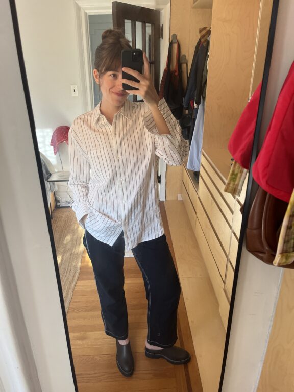 Person taking a mirror selfie in a bedroom, wearing a white, striped button-down shirt, dark pants, and black shoes. One hand is in their pocket, and the other holds the phone.