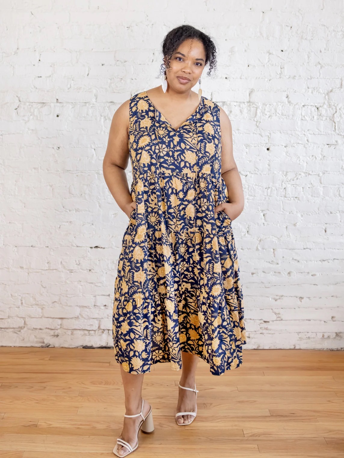 A woman stands indoors on a wooden floor, wearing a sleeveless navy and tan floral dress and white sandals, posing with hands in pockets against a white brick wall.