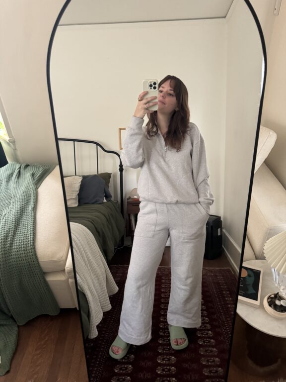 A woman stands in front of a full-length mirror taking a selfie, wearing a light gray loungewear set and slippers in a neatly furnished bedroom.