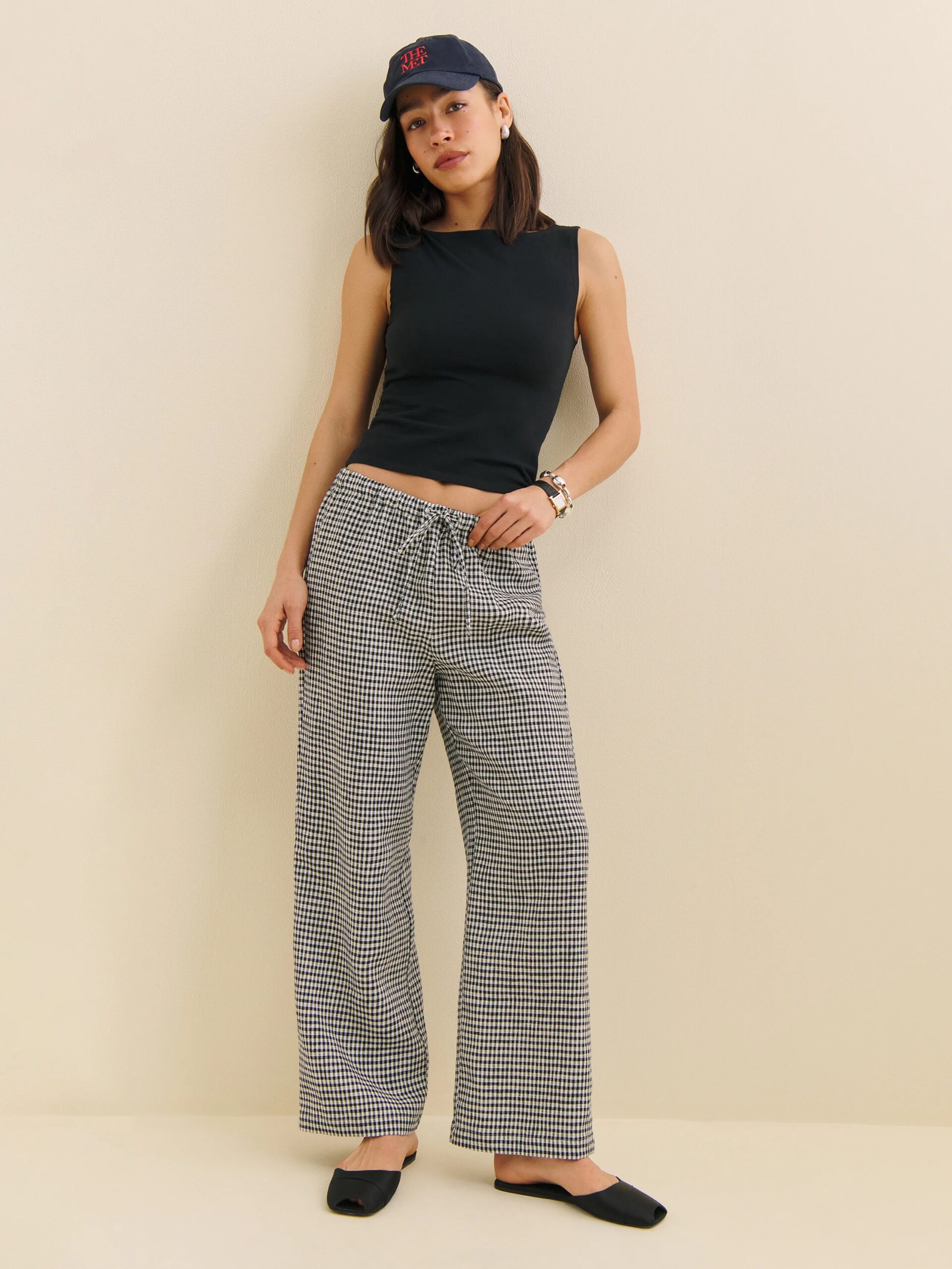 A woman stands against a plain wall wearing a black sleeveless top, checkered wide-leg pants, black shoes, a black baseball cap, and a bracelet.