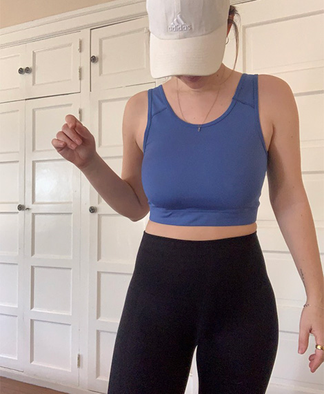 Person wearing a blue crop top, black leggings, and a white Adidas cap stands indoors in front of white paneled cabinets.
