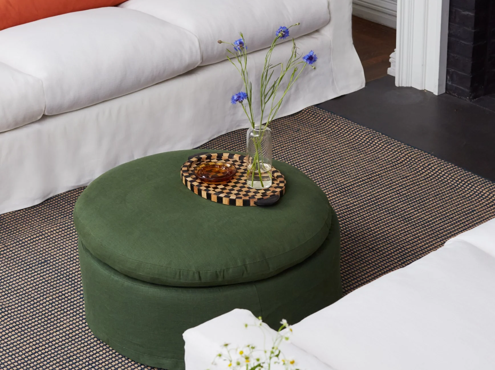 A round green ottoman with a checkered tray, glass vase with blue flowers, and a small bowl sits on a patterned rug between two white sofas.