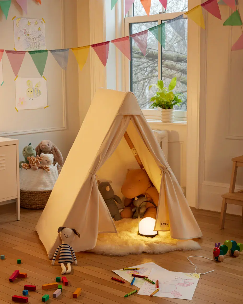 A children’s playroom with a small fabric teepee, stuffed animals inside, a glowing lamp, colorful bunting, drawings on the wall, toys on the floor, and a window in the background.