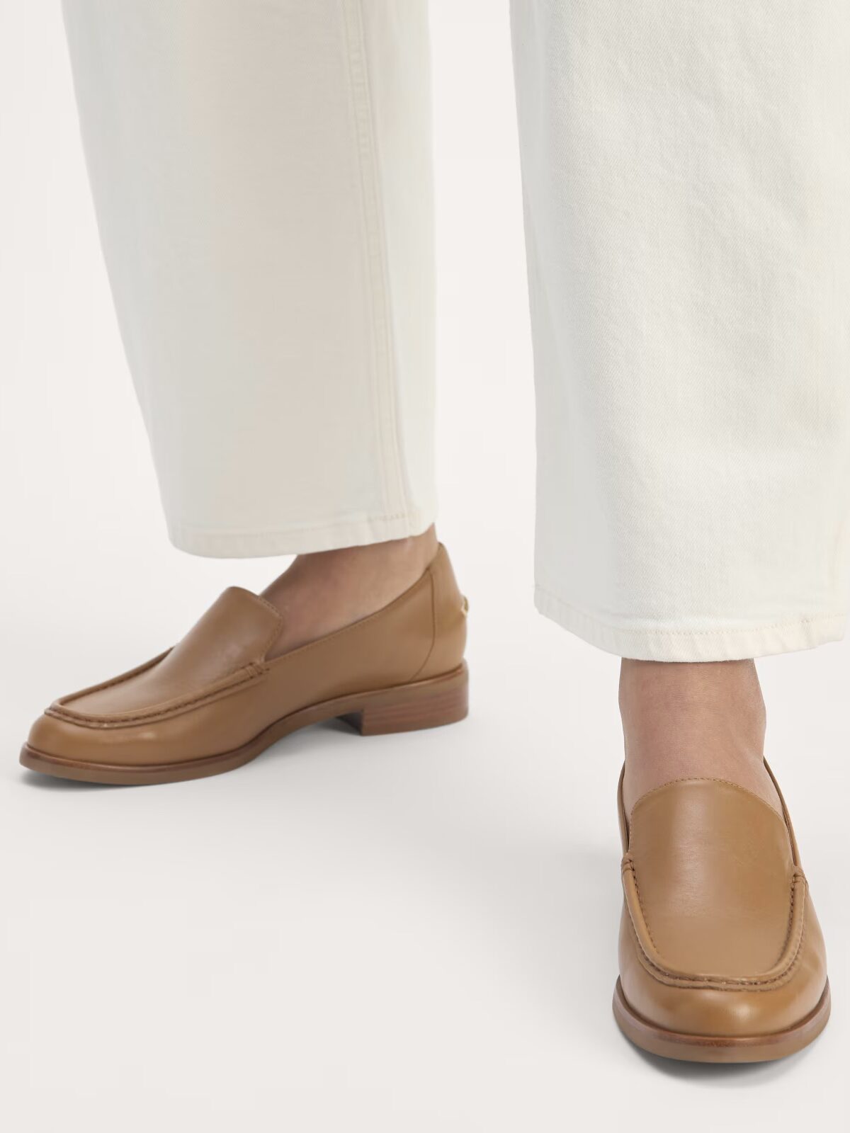 A person wearing white pants and light brown leather loafers stands on a light background.