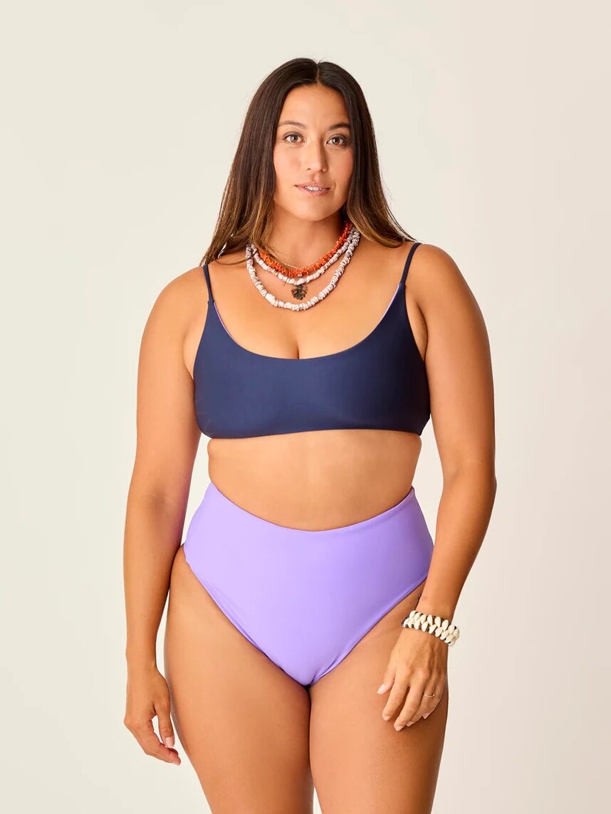 Woman wearing a navy bikini top, high-waisted lavender bikini bottoms, layered necklaces, and a bracelet, standing against a plain light background.