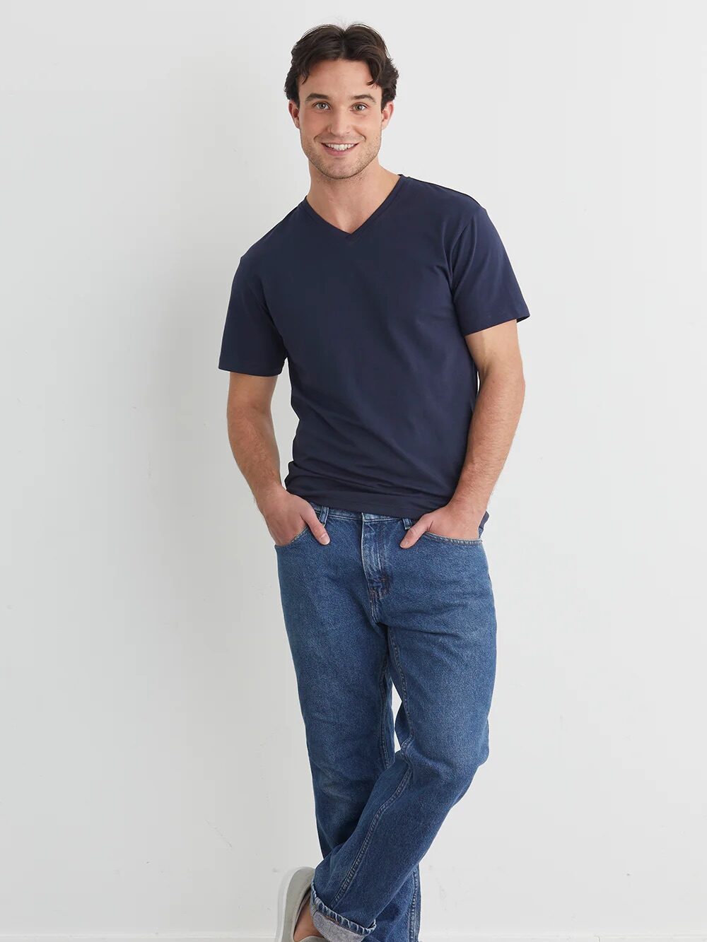 A man stands against a white wall wearing a navy V-neck T-shirt, blue jeans, and gray slip-on shoes, with one foot crossed over the other and hands in his pockets.