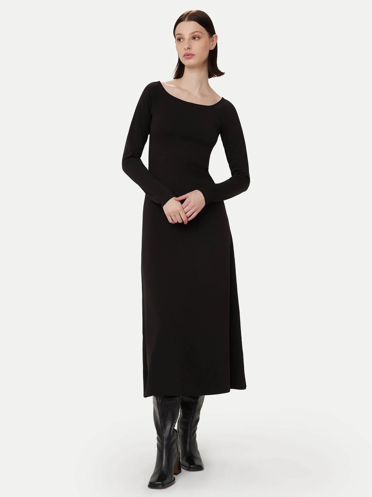 A woman stands wearing a long-sleeved, ankle-length black dress and black boots against a plain light background.