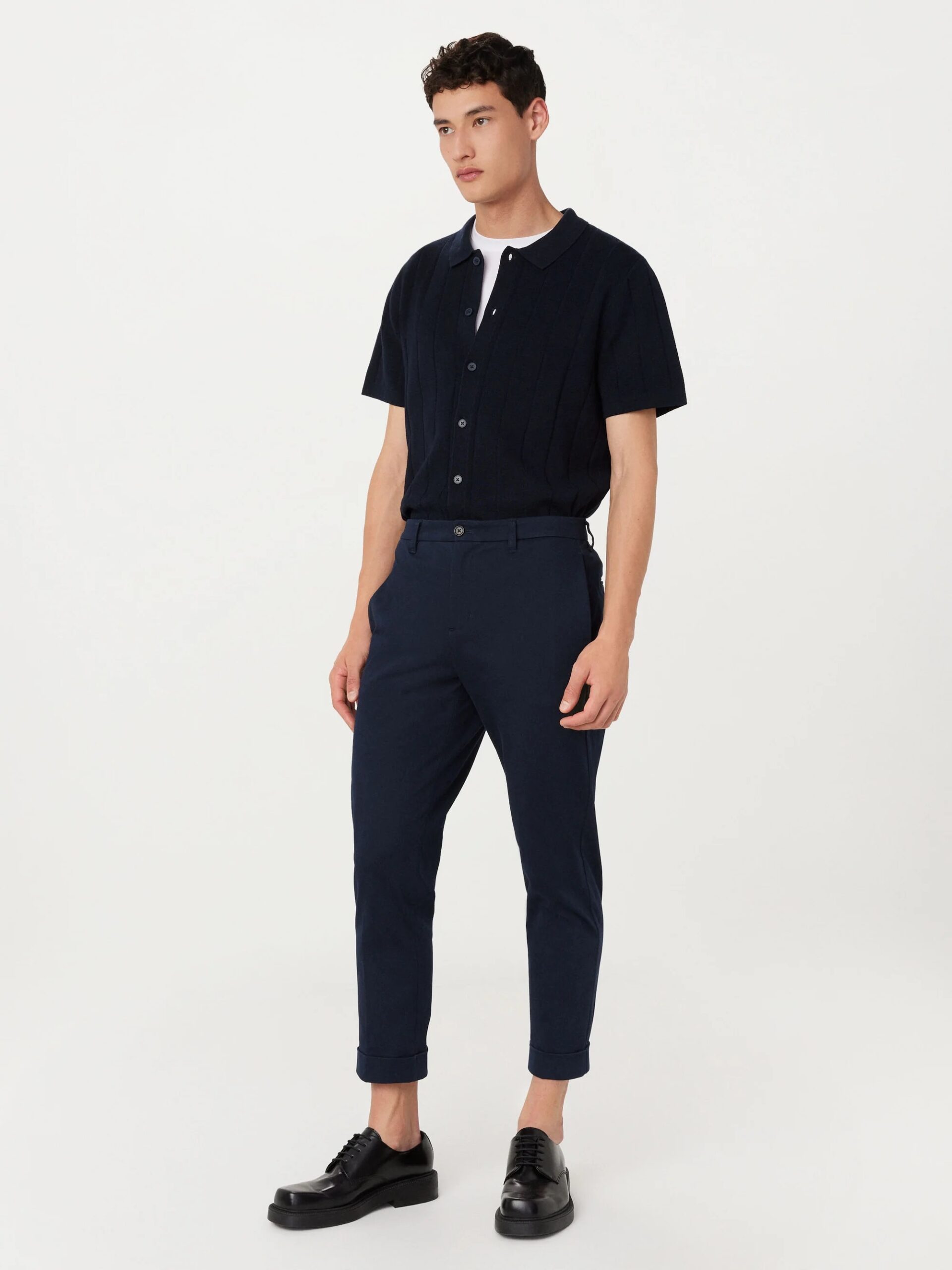A man stands against a plain background wearing a black short-sleeve shirt over a white tee, navy blue pants, and black shoes.
