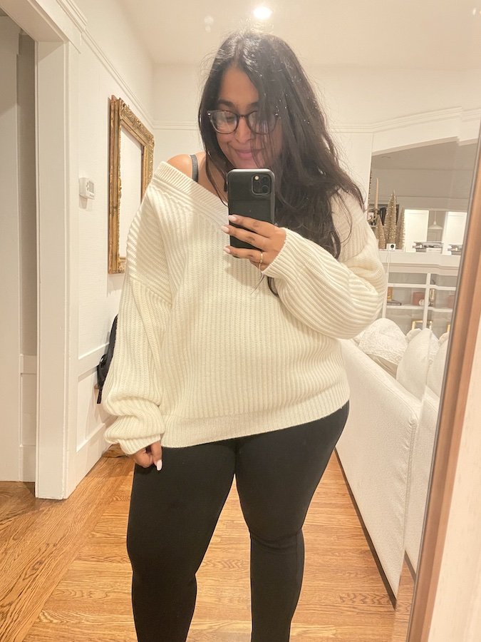 Woman taking a mirror selfie in a well-lit room, wearing a white oversized sweater and black leggings.