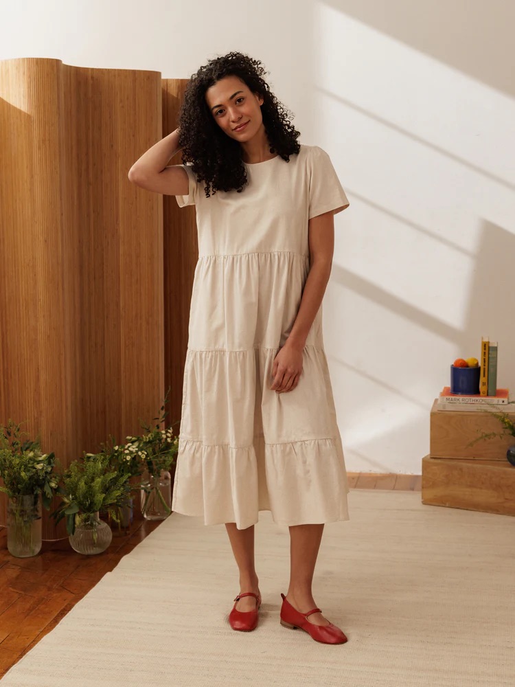 A woman stands indoors on a light rug, wearing a short-sleeve cream tiered dress and red shoes, with plants and stacked boxes in the background.
