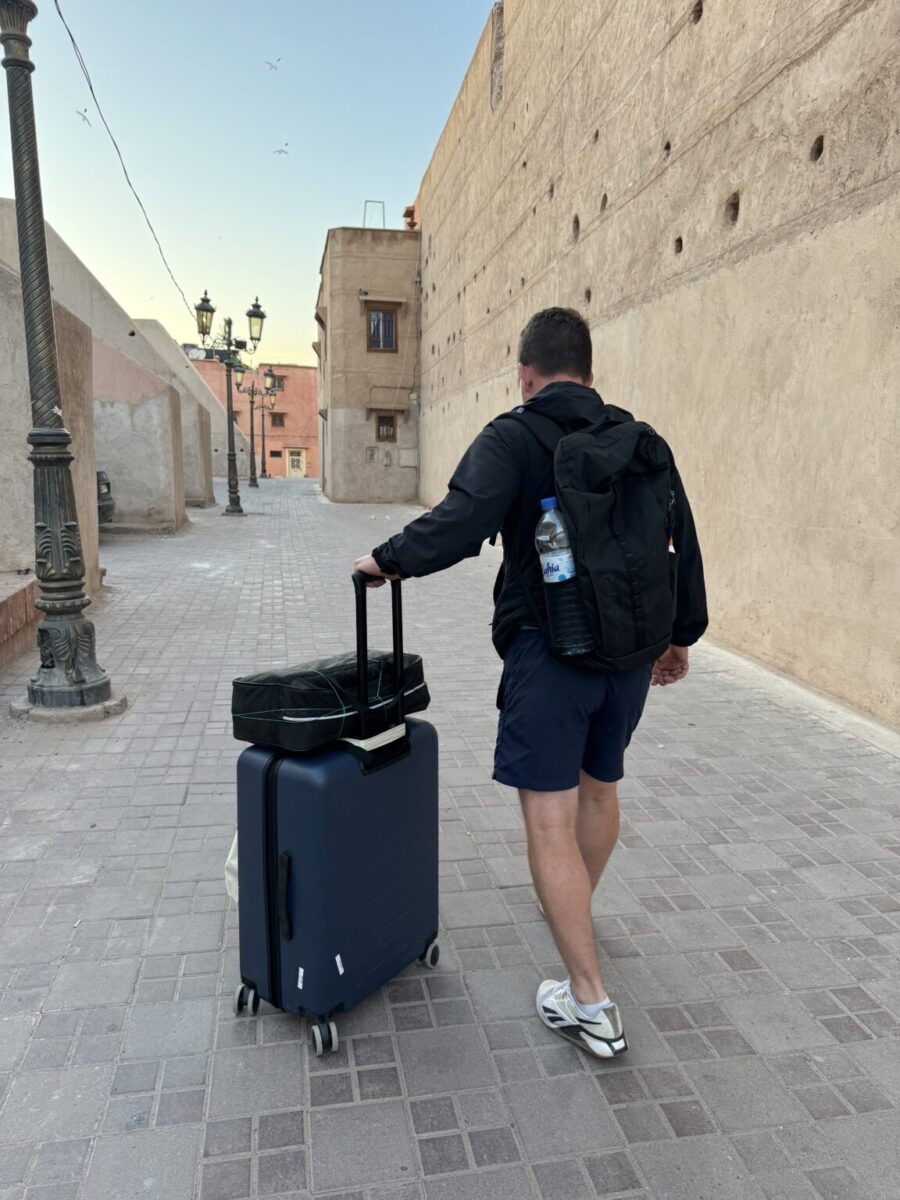 A man wearing shorts and a backpack pulls a suitcase down a narrow, empty cobblestone street with beige walls.