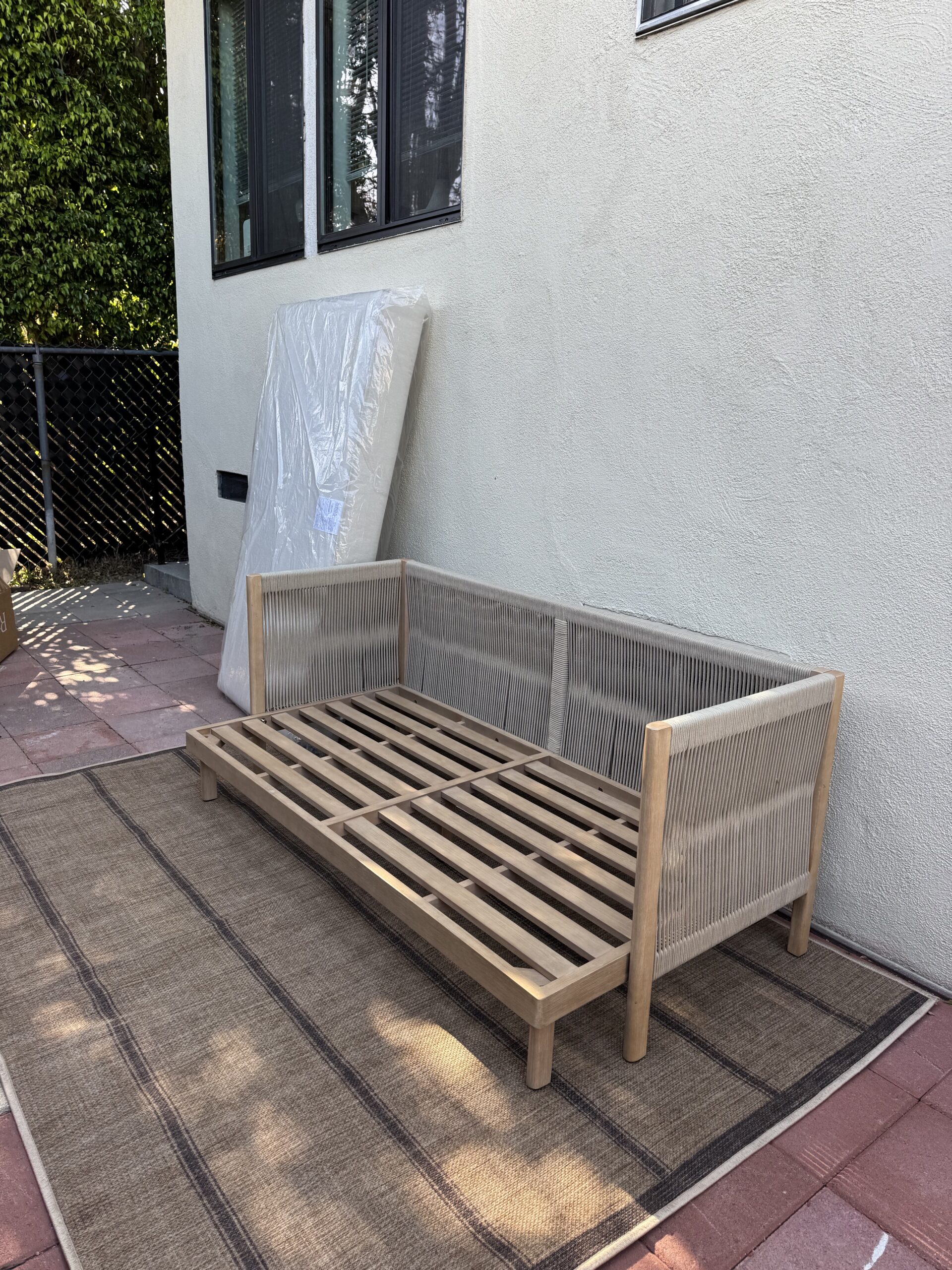 A wooden outdoor sofa frame with rope details sits on a woven rug next to a white wall and a wrapped cushion, on a tile patio.