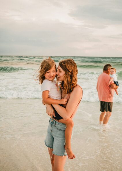 A woman smiles while holding a young girl at the beach; a man in the background holds another child near the water.