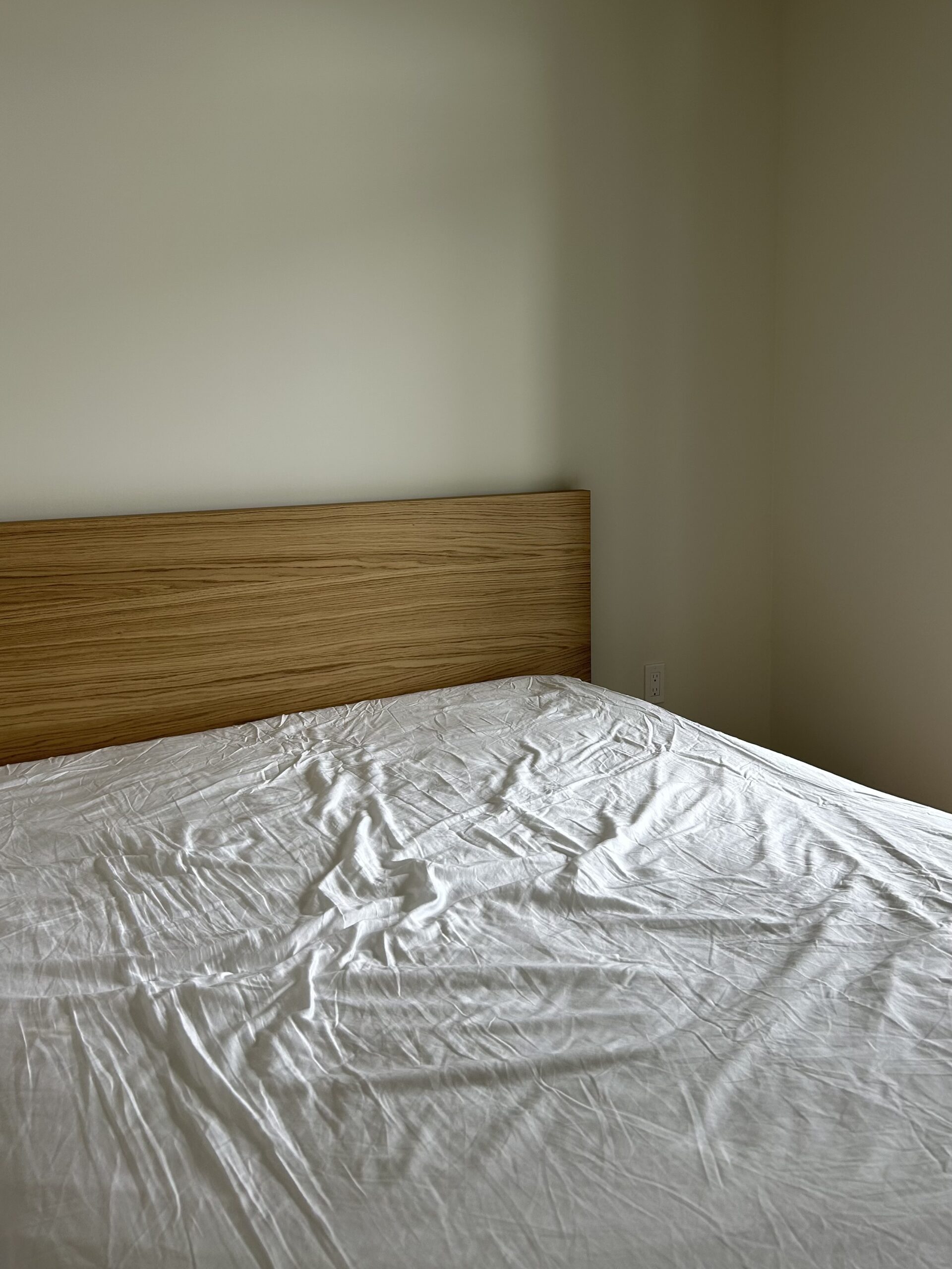 A simple bed with a wooden headboard and white wrinkled sheets in a bare, light-colored room.
