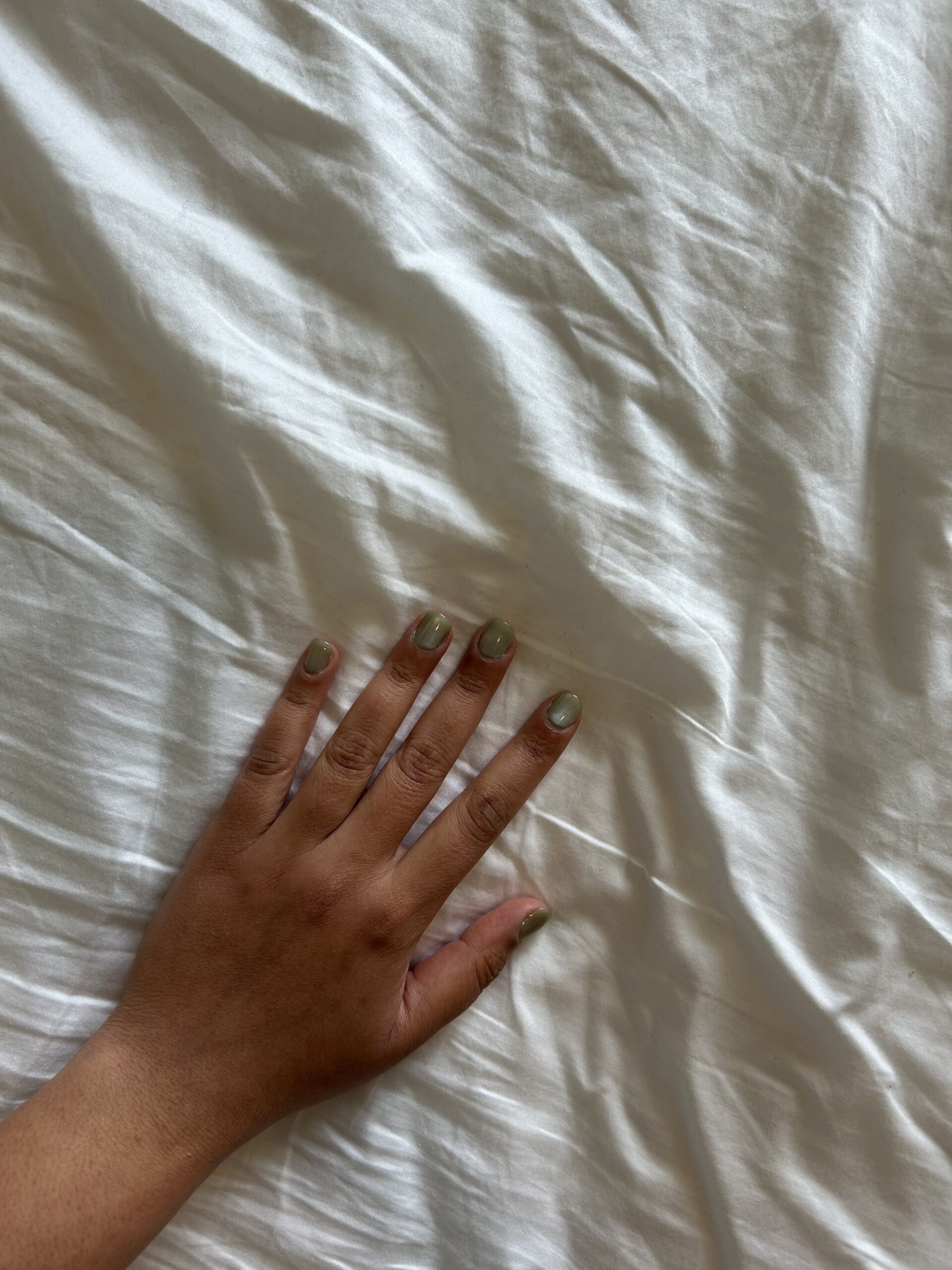 A left hand with light green painted nails rests on a white, wrinkled sheet.