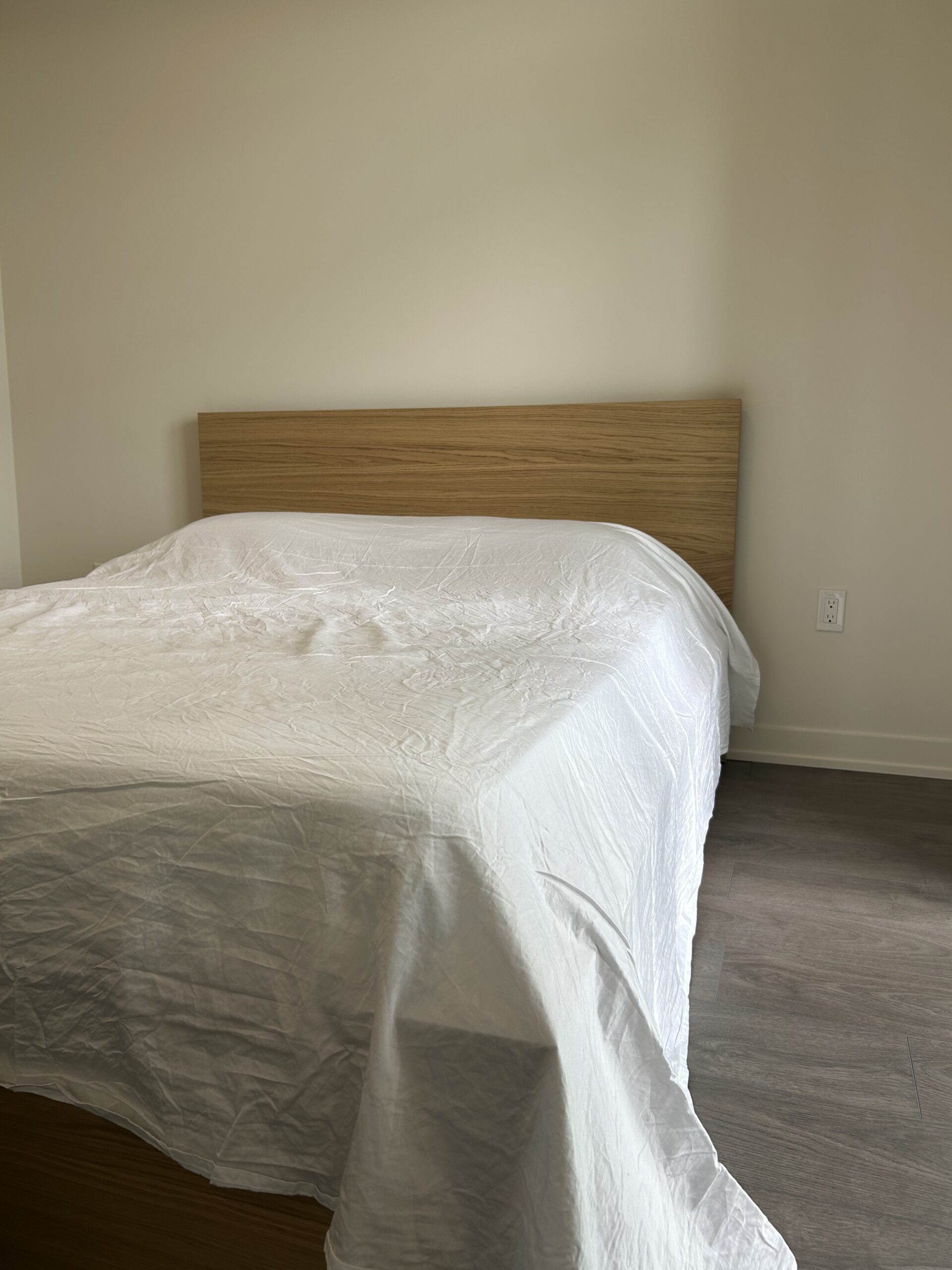 A bed with a wooden headboard is covered with a white sheet in a minimally decorated room with light-colored walls and gray flooring.