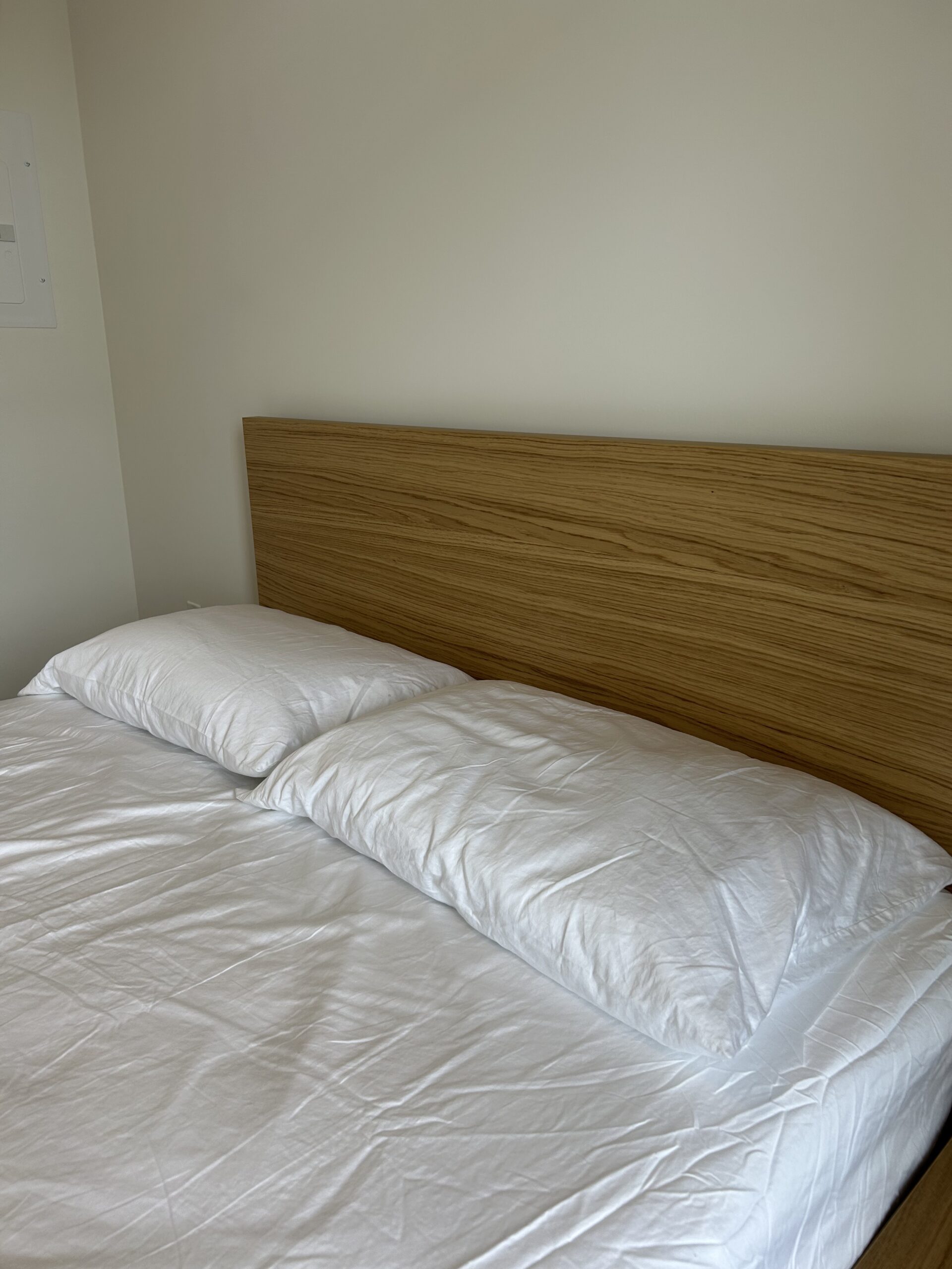 A neatly made bed with white sheets and two pillows, featuring a wooden headboard against a plain light-colored wall.
