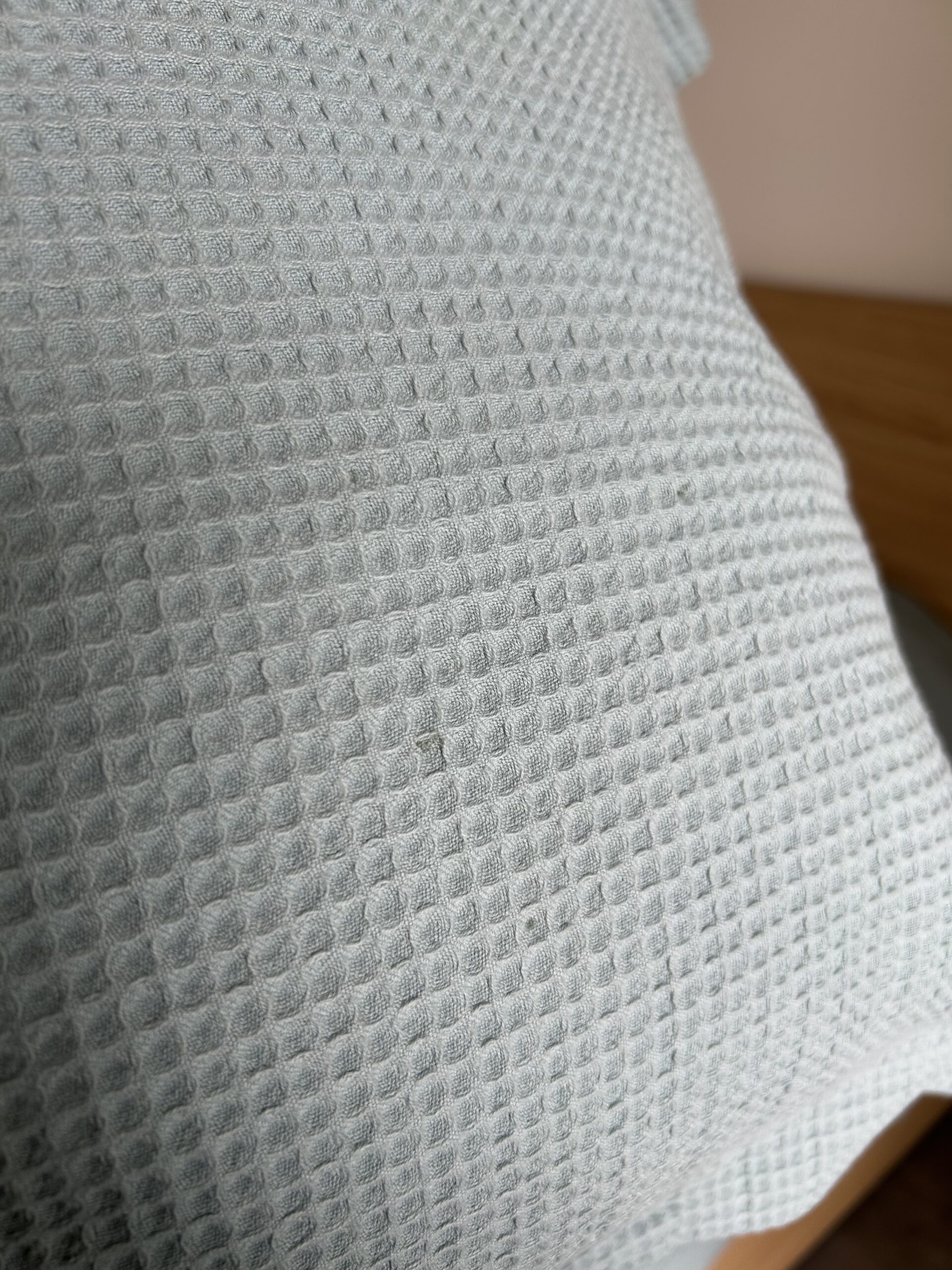 Close-up of a light gray textured fabric pillow with a waffle weave pattern, positioned against a wooden surface.