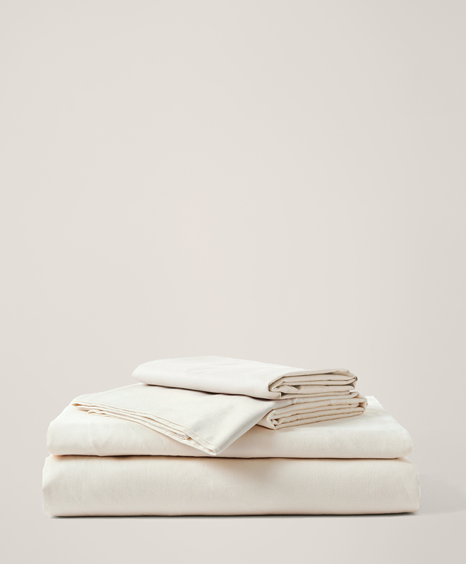 A neatly folded stack of cream-colored bed sheets and pillowcases is arranged against a plain, light background.