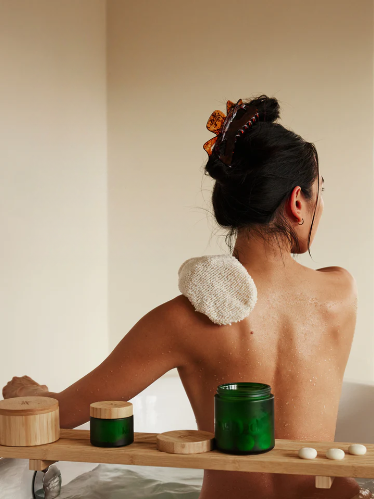 A woman with a hair clip exfoliates her back in a bath using a textured sponge. Skincare jars and containers are arranged on a wooden tray nearby.