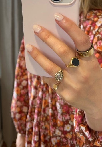 A hand with manicured nails holds a phone, wearing four gold rings with various designs, against a background of a floral-patterned dress.