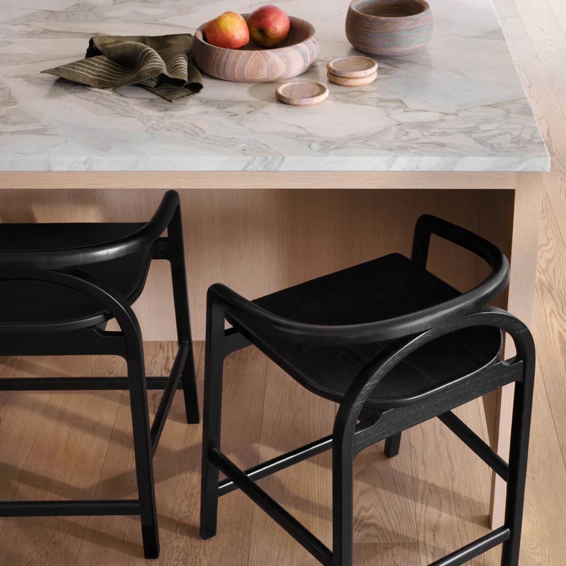 Two black stools are placed by a marble countertop with a wooden bowl of apples, a cloth napkin, and two small lidded containers on top.